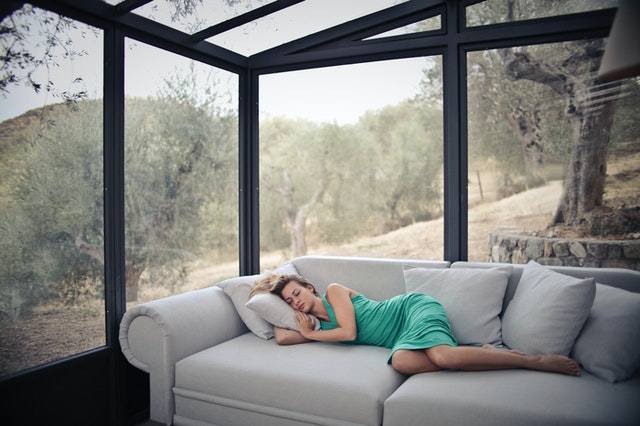 Žena v zelených šatách spí na gauči v miestnosti so sklenenými stenami 